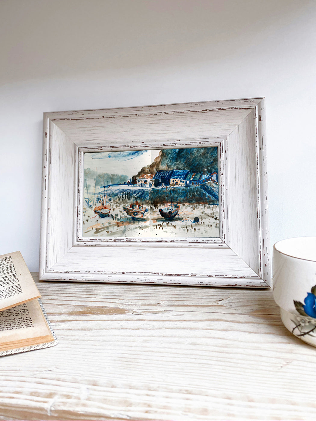 Framed painting of a coastal scene on a wooden surface with a book and mug.